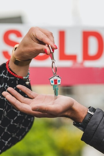 Real Estate Agent handing new home owner a key to their new house. A "Sold" sign is in the background.