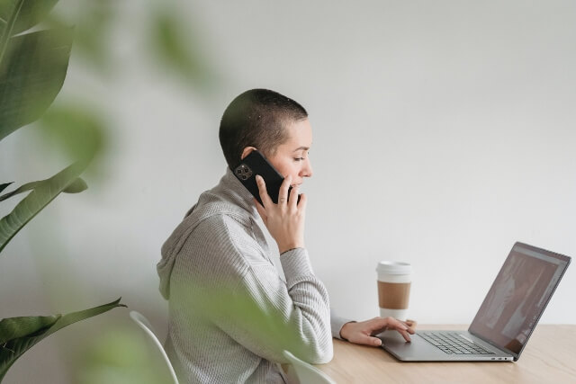 Woman working on computer and talking on phone. A cup of coffee is sitting next to her and a green plant is in the foreground.